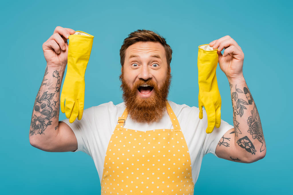 zdumiony wytatuowany mężczyzna w fartuchu pokazując żółte gumowe rękawiczki patrząc na aparat odizolowany na niebiesko - Zdjęcie, obraz