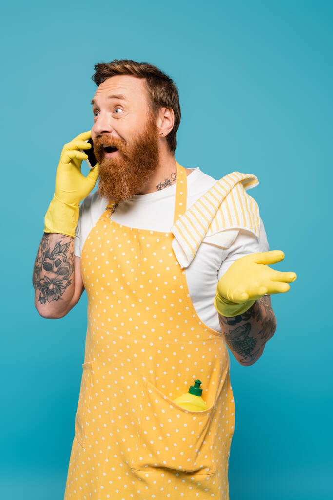 hombre barbudo excitado en delantal de lunares hablando en el teléfono inteligente y apuntando con la mano aislada en azul - Foto, imagen