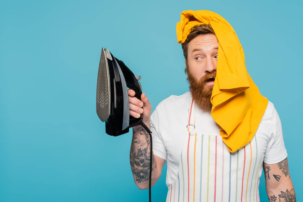 bearded man with yellow garment on head looking at electric iron isolated on blue - Foto, immagini
