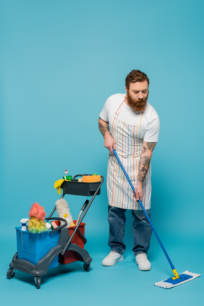 full length of serious bearded man with mop cleaning floor near cart with cleaning supplies on blue background - Foto, immagini