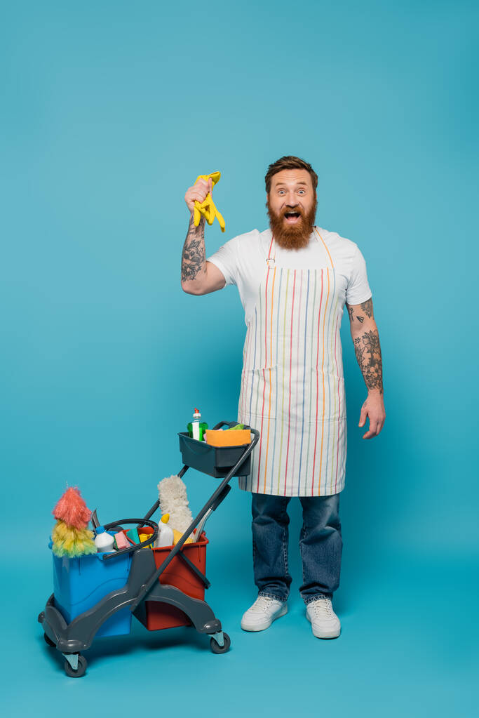 amazed bearded man in apron holding rubber gloves near cart with cleaning supplies on blue background - Fotoğraf, Görsel