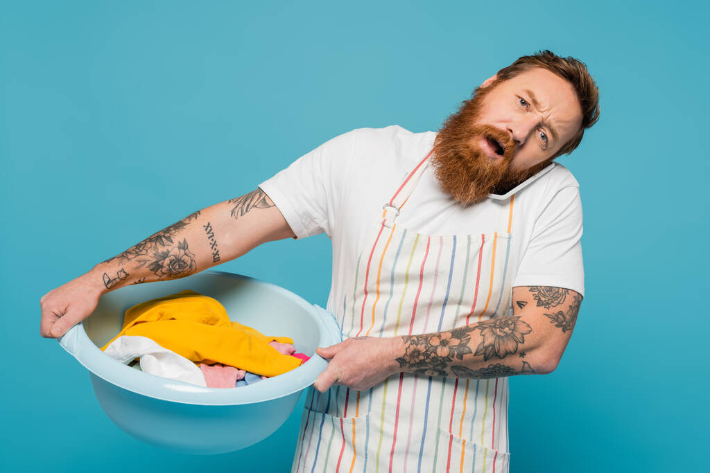exhausted tattooed man talking on mobile phone while holding laundry bowl isolated on blue - Fotoğraf, Görsel