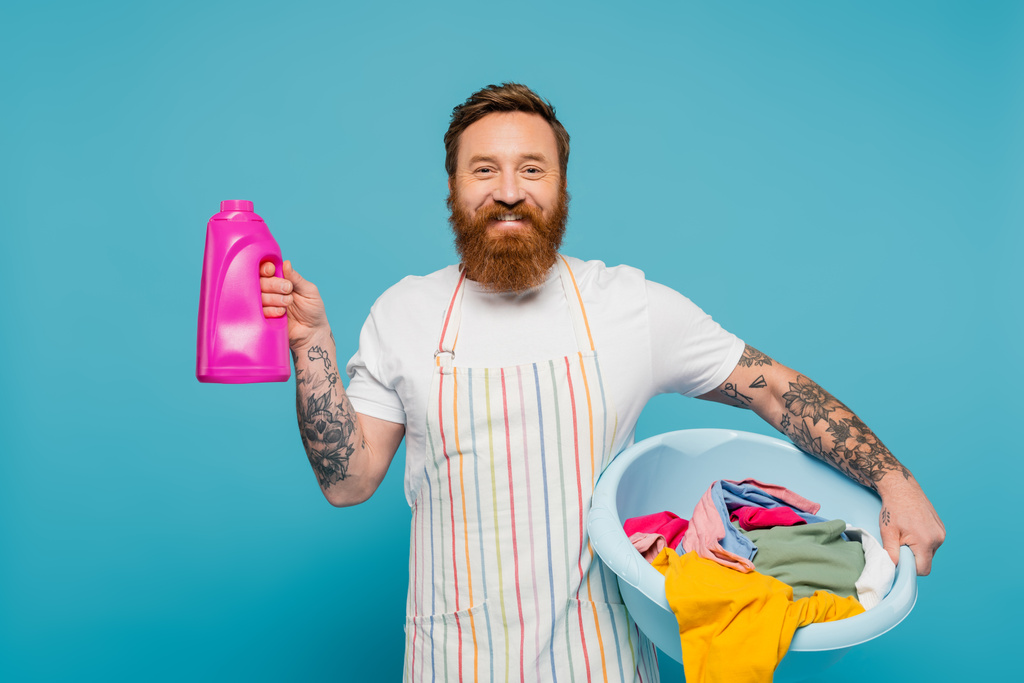 positive tattooed man with washing gel and laundry bowl looking at camera isolated on blue - Foto, Imagem