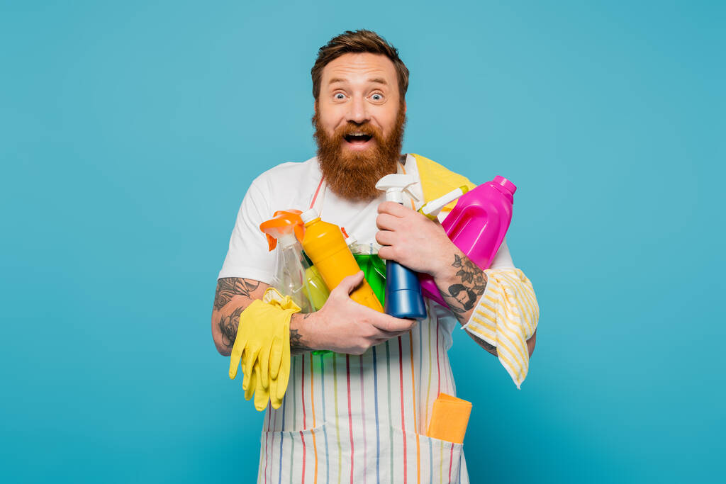 изумленный и счастливый человек, держащий в руках различные моющие средства и резиновые перчатки, глядя на камеру, изолированную на голубом - Фото, изображение