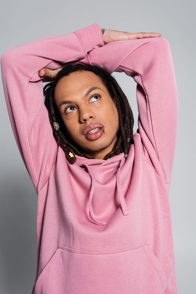 tattooed multiracial man in pink hoodie looking up while thinking on grey  - Foto, imagen