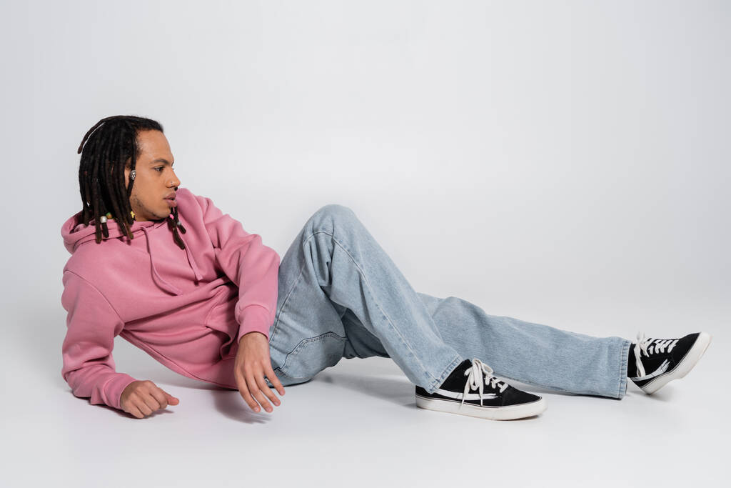 side view of multiracial and pierced man with dreadlocks sitting on grey background  - Foto, imagen
