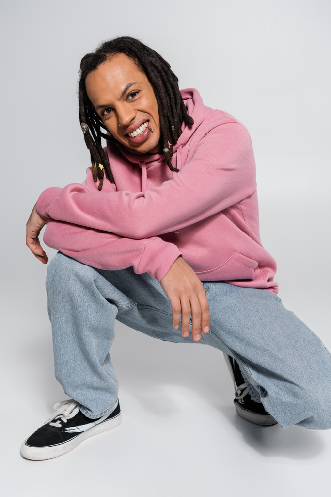 full length of positive and multiracial man with dreadlocks posing on grey  - Foto, afbeelding