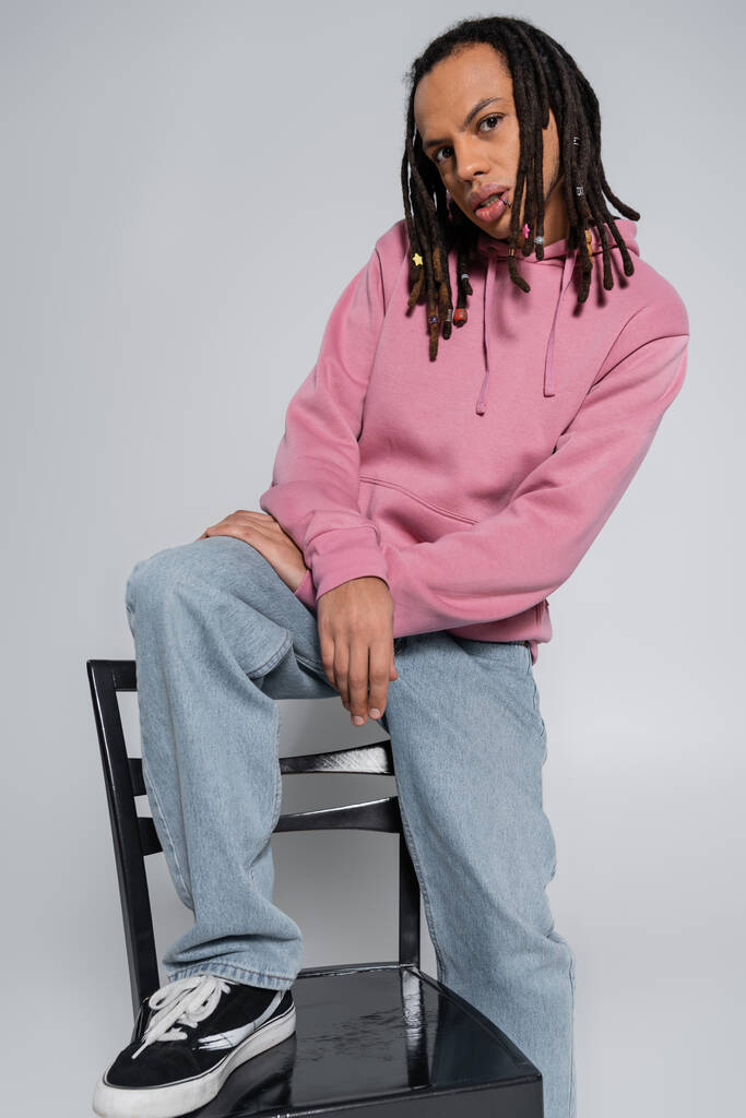 multiracial and pierced man with dreadlocks looking at camera while sitting on chair back isolated on grey  - Foto, immagini