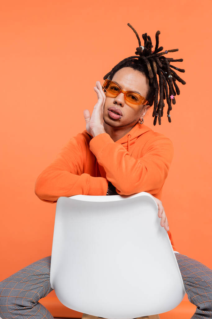 multiracial man with dreadlocks and sunglasses sitting on chair isolated on coral  - Foto, Imagem
