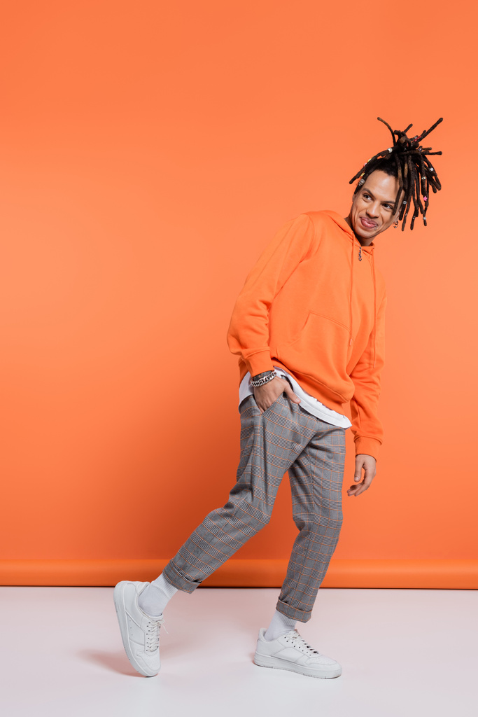 full length of smiling multiracial man with dreadlocks standing with hand in pocket on coral background  - Foto, Imagen