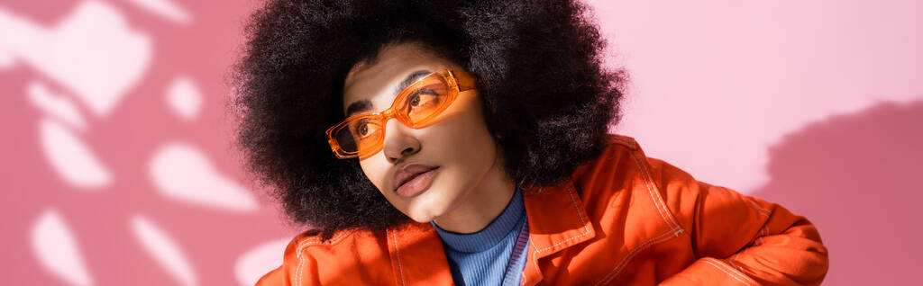femme afro-américaine bouclée dans des lunettes de soleil orange élégant regardant loin sur rose, bannière   - Photo, image