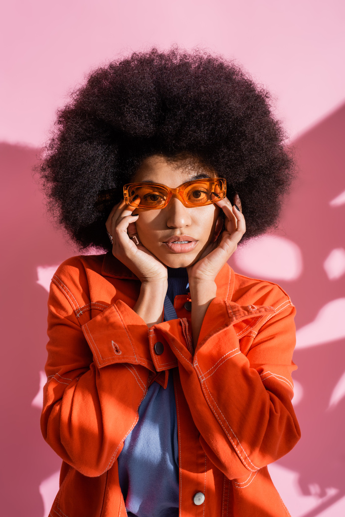молодая африканская американка в стильных оранжевых солнцезащитных очках и модном наряде позирует на розовом  - Фото, изображение
