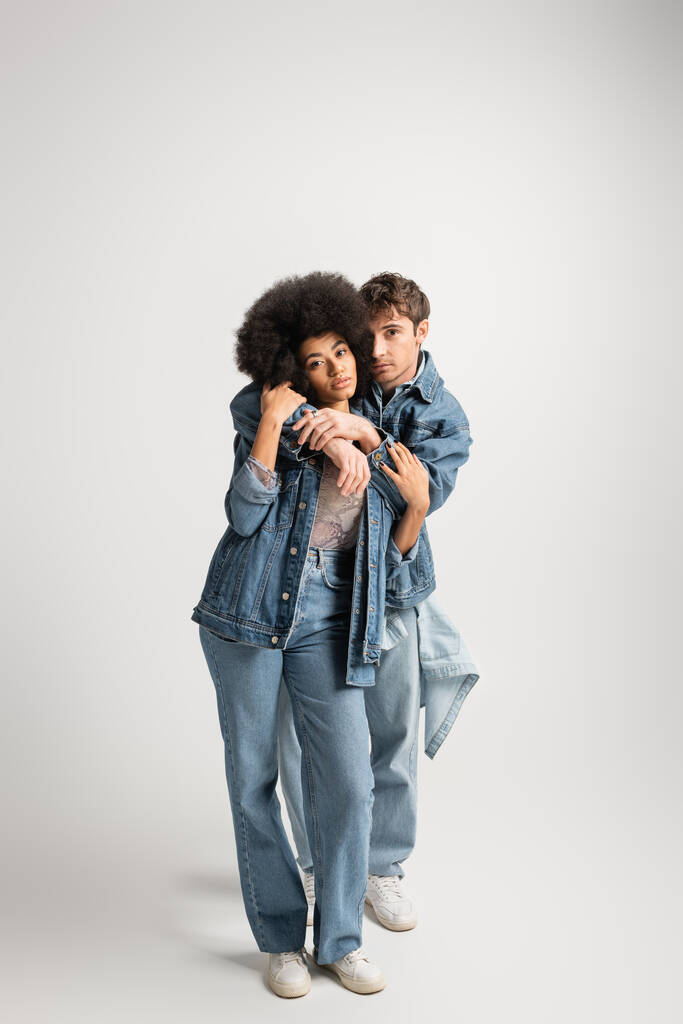 full length of young man in denim outfit hugging curly african american woman on grey  - Foto, Imagem