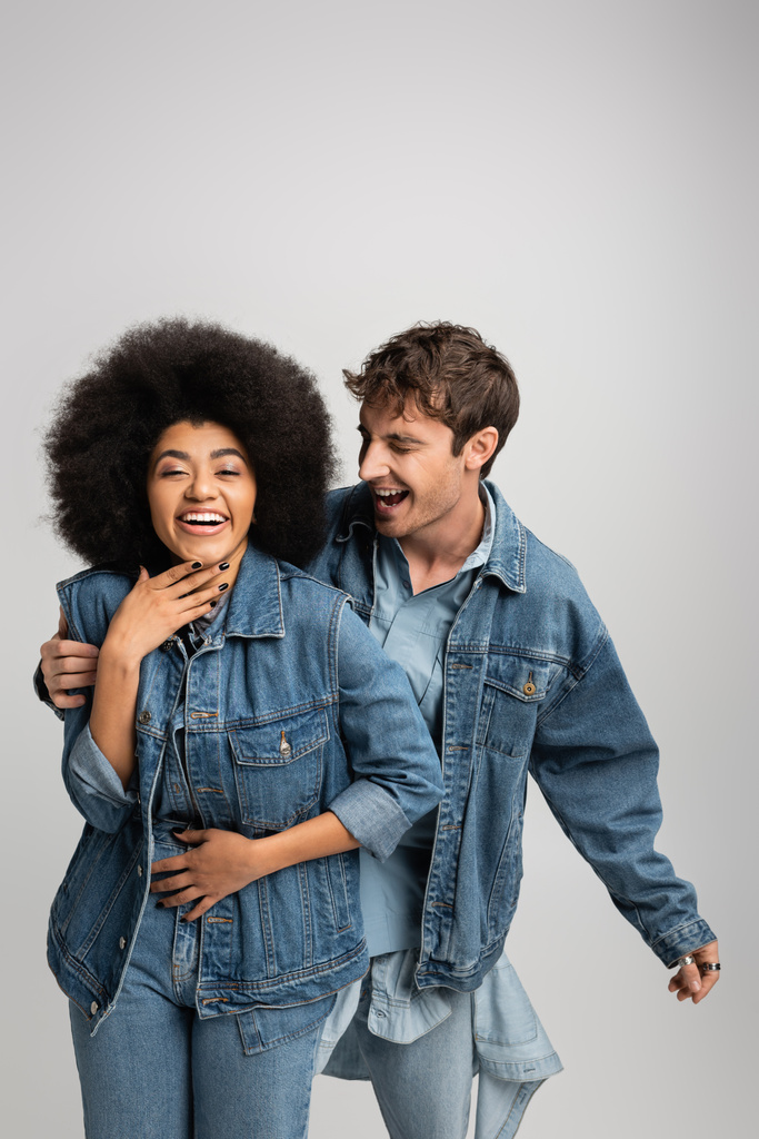 feliz hombre y mujer multiétnicos en traje de mezclilla riendo aislado en gris  - Foto, imagen