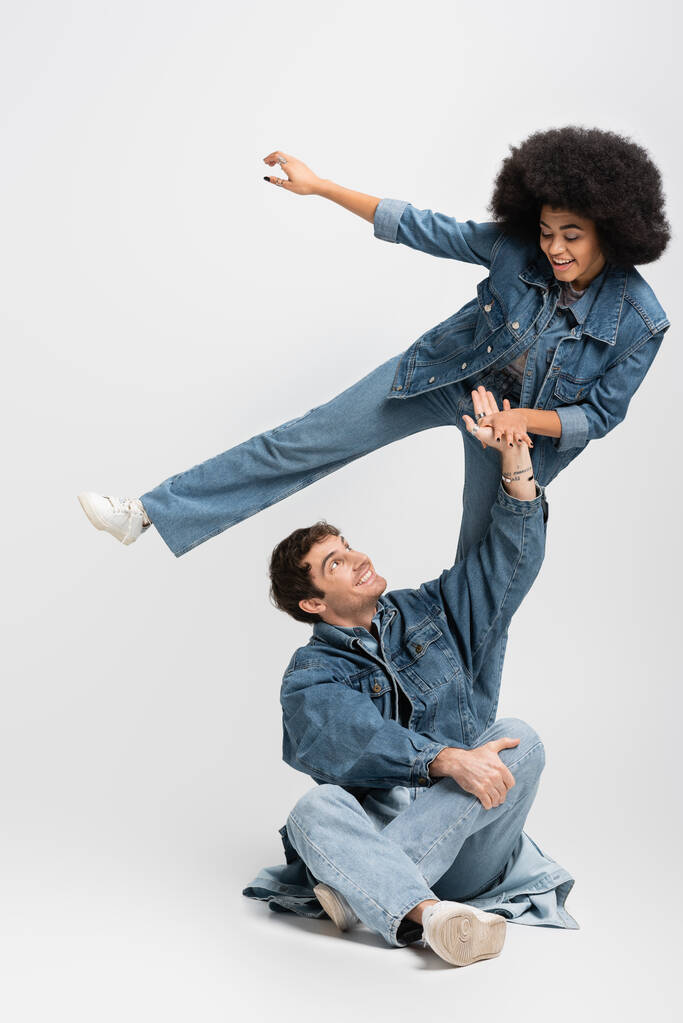 повна довжина щасливої афроамериканки позує біля веселого чоловіка в джинсовому вбранні і кросівки, сидячи на сірому
 - Фото, зображення
