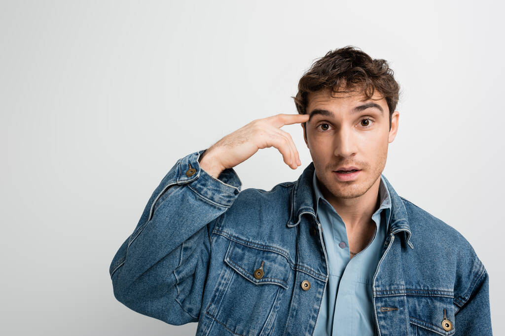 overwhelmed man in denim jacket pointing with finger at head and looking at camera isolated on grey  - Foto, Imagem