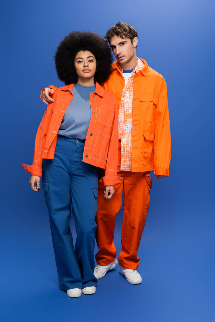 Stylish man hugging african american woman in orange jacket on blue background - Zdjęcie, obraz
