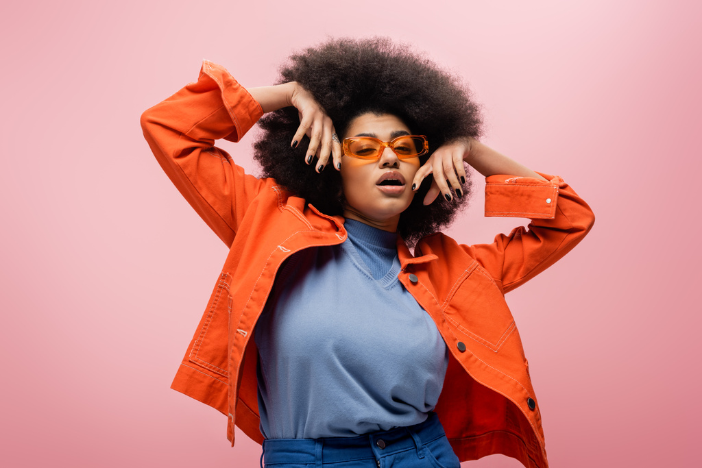 Модная африканская американская модель в оранжевой куртке и солнечных очках позирует изолированно на розовом  - Фото, изображение