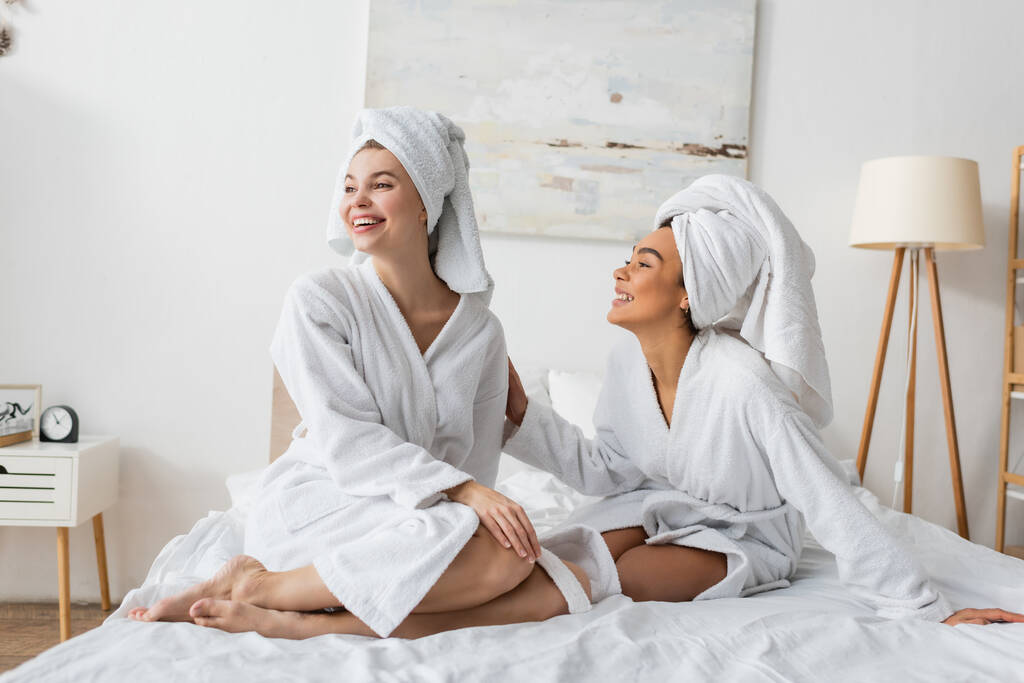 feliz jovem mulher em terry roupão de banho e toalha olhando para longe perto sorrindo amigo afro-americano na cama - Foto, Imagem