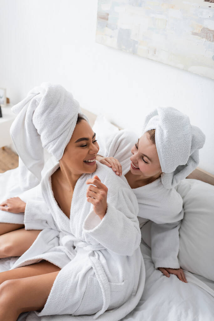 vista de ángulo alto de las mujeres jóvenes multiétnicas en toallas blancas y albornoces sonrientes entre sí en la cama - Foto, imagen