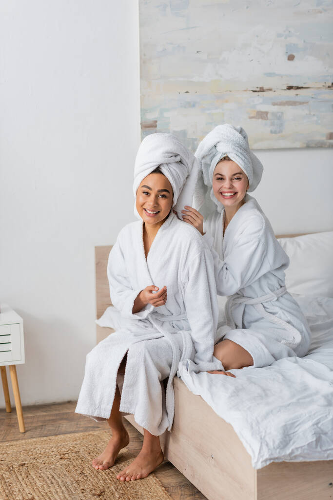 szczęśliwy międzyrasowe kobiety w biały miękkie szaty i ręczniki patrząc w aparat podczas siedzi na łóżku - Zdjęcie, obraz