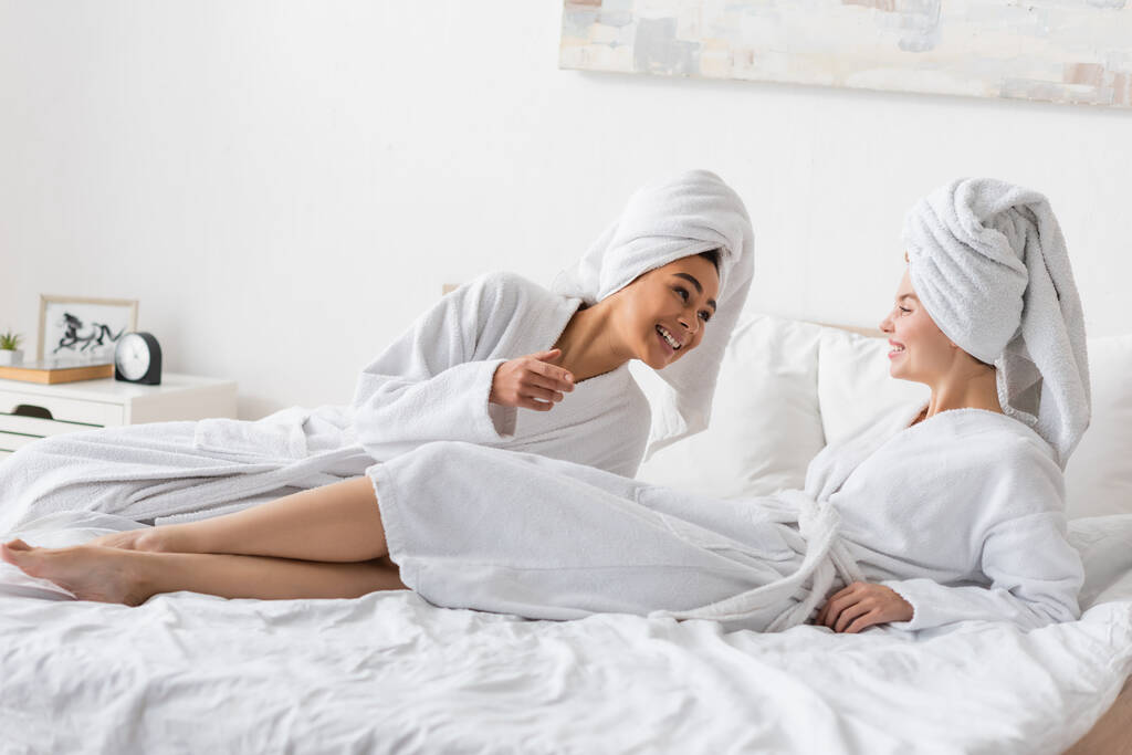 Χαρούμενη Αφρικανή Αμερικανίδα που μιλάει σε μια όμορφη φίλη ξαπλωμένη στο κρεβάτι με λευκό μπουρνούζι και πετσέτα.  - Φωτογραφία, εικόνα