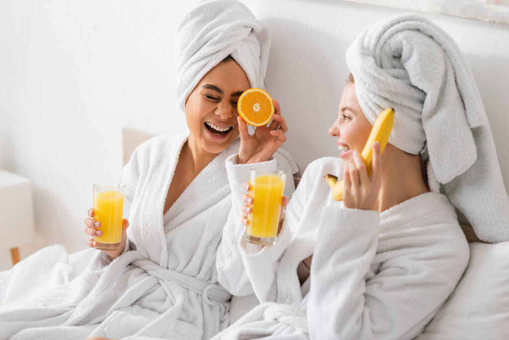 young multiethnic women in white robes and towels having fun with ripe fruits in bedroom - 写真・画像