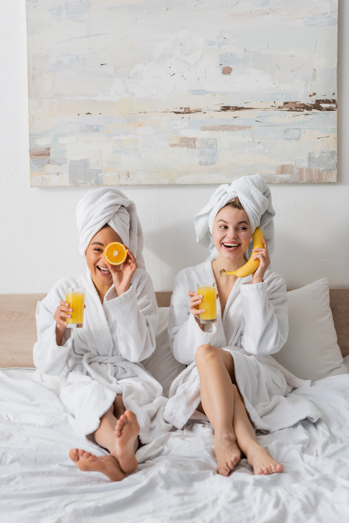 full length of cheerful interracial women with fruits and orange juice having fun while sitting on bed - Foto, immagini