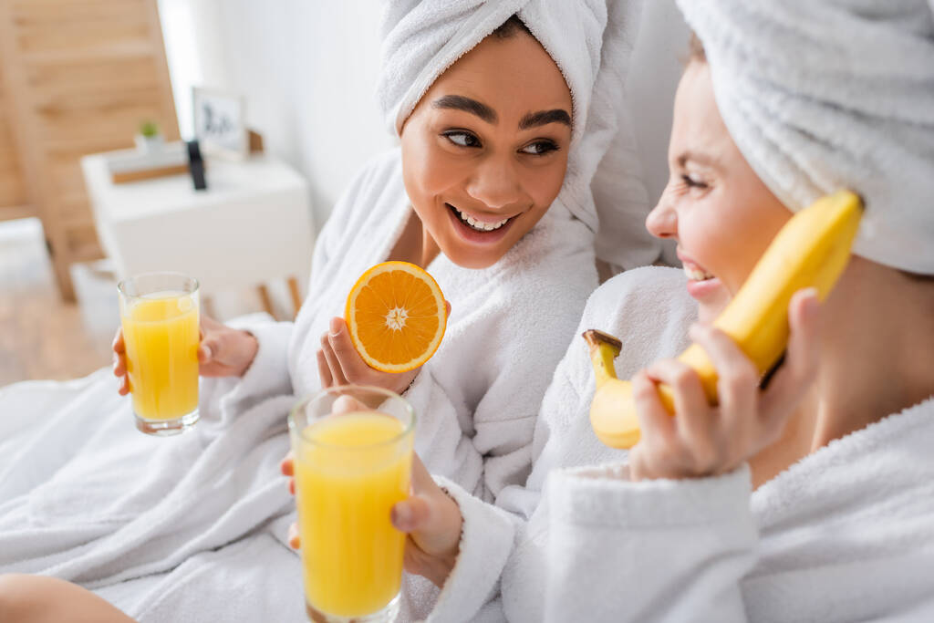 happy african american woman holding ripe orange and juice near blurred friend having fun in bedroom - Photo, Image