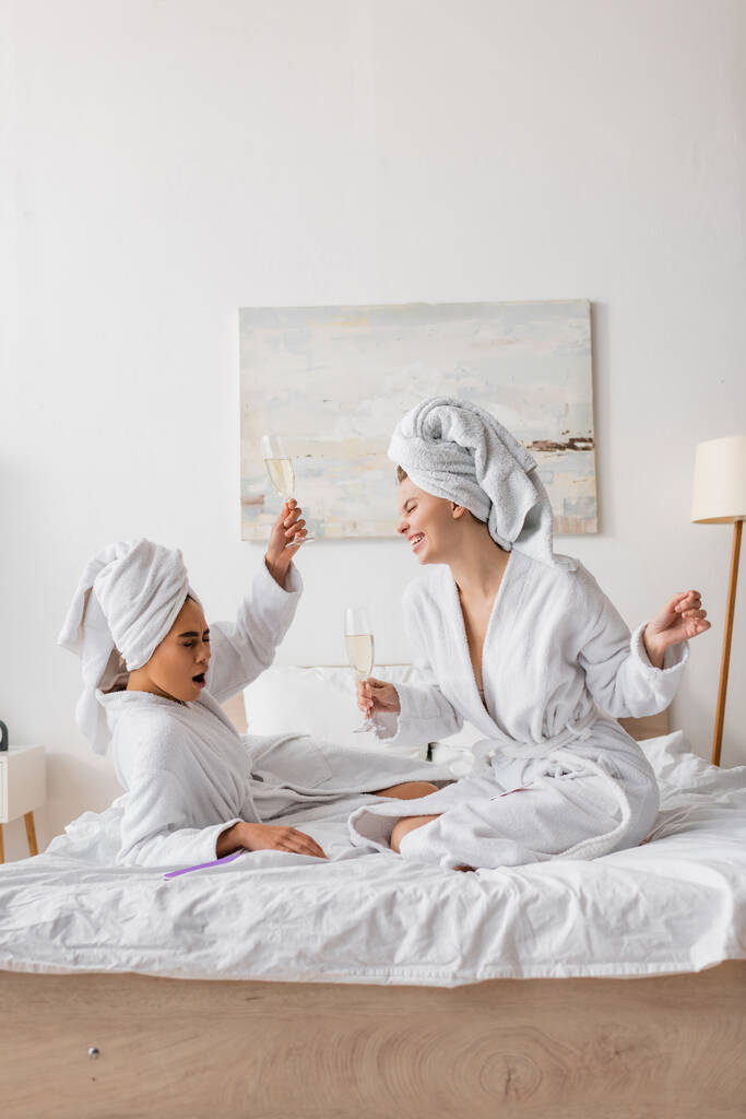 opgewonden Afrikaans amerikaanse vrouw toasten met champagne in de buurt gelukkig vriend in witte badjas en handdoek in slaapkamer - Foto, afbeelding