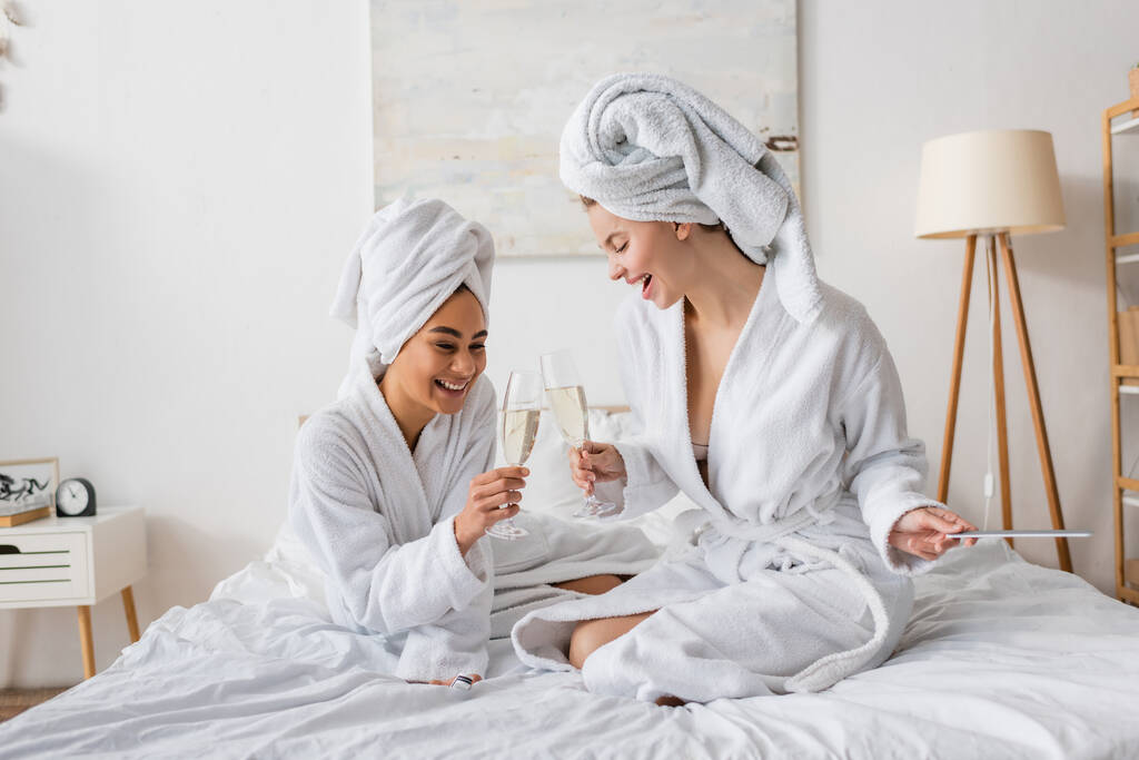 cheerful interracial women in white soft bathrobes and towels clinking champagne glasses on bed  - Photo, Image