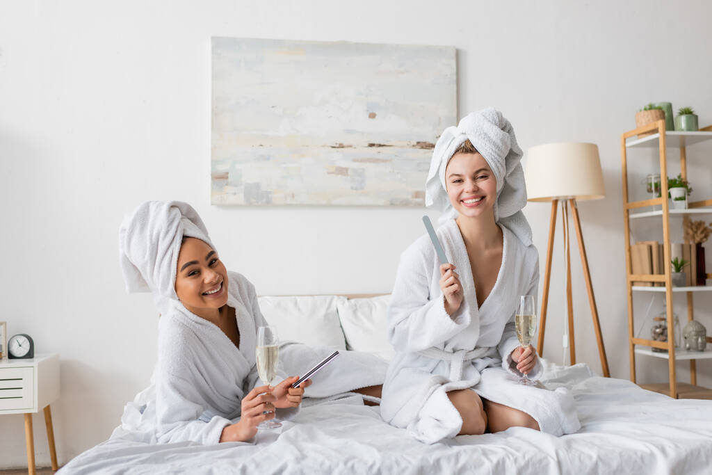 cheerful interracial women with champagne glasses and nail files looking at camera on bed - Photo, Image