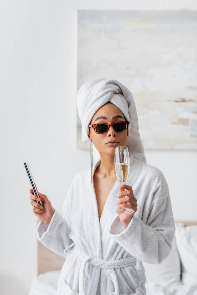 Beyaz bornozlu ve şık güneş gözlüklü genç Afrikalı Amerikalı kadın elinde şampanya bardağı ve tırnak törpüsüyle yatak odasında. - Fotoğraf, Görsel