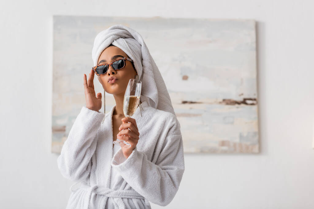 Terry bornoz ve havlu giymiş Afro-Amerikan kadın dudak bükerek ve yatak odasında şampanya tutarak son moda güneş gözlüklerini ayarlıyor. - Fotoğraf, Görsel