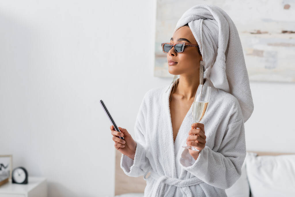 young african american woman in soft terry bathrobe and sunglasses looking away while holding nail fail and champagne in bedroom - Photo, Image