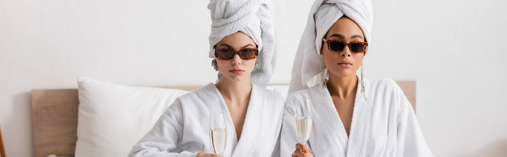 jonge multi-etnische vrouwen in witte gewaden en een zonnebril kijken naar de camera terwijl het houden van champagne glazen in de slaapkamer, banner - Foto, afbeelding