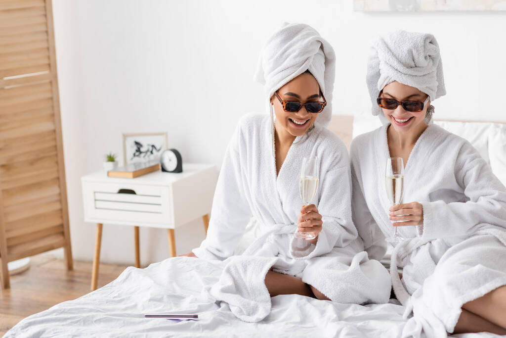 pleased multiethnic women in soft bathrobes and sunglasses sitting with champagne glasses on bed - Foto, afbeelding