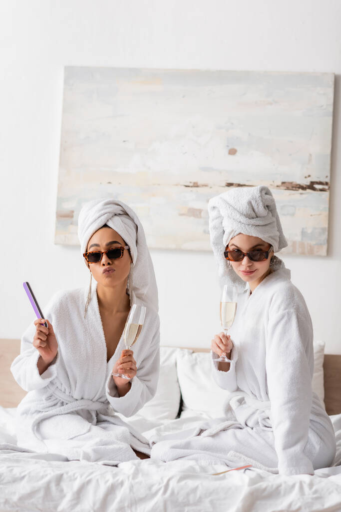 young interracial women in white bathrobes and stylish sunglasses posing with champagne glasses and nail file on bed - Photo, Image