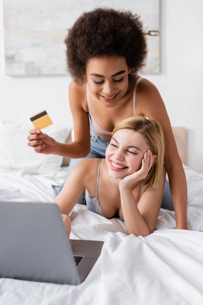 Genç Afrikalı Amerikalı bir kadın kredi kartı tutuyor. Yanında mutlu sarışın bir kadın. Yatak odasında bulanık dizüstü bilgisayara bakıyor. - Fotoğraf, Görsel