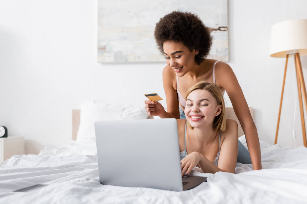happy african american woman holding credit card near smiling friend using laptop on bed - Fotó, kép