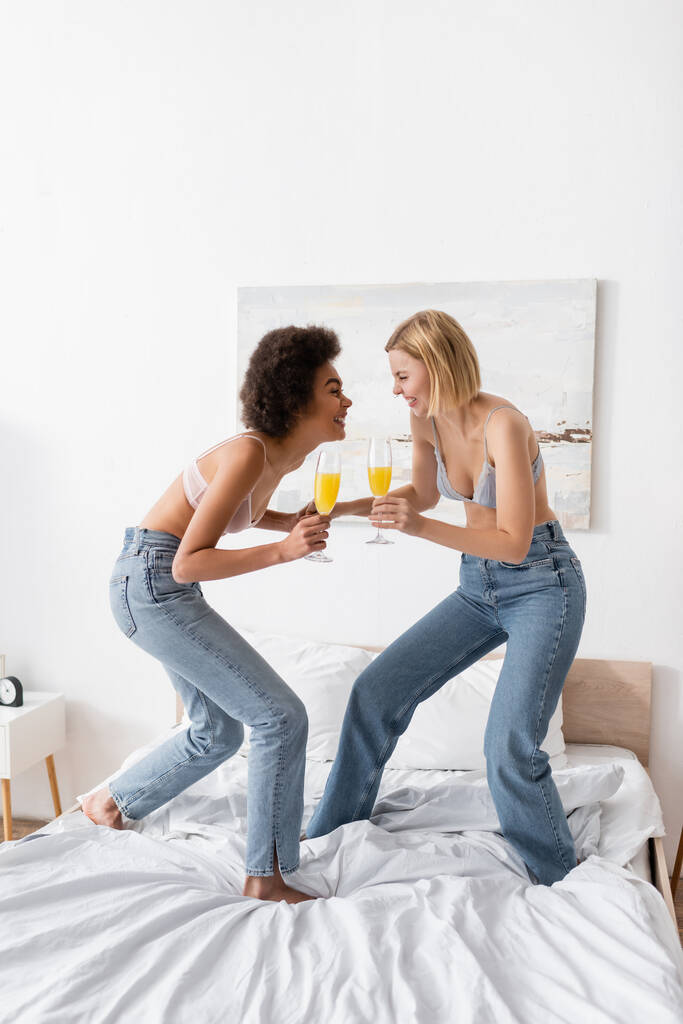full length of excited multiethnic women holding champagne glasses with cocktails while having fun on bed at home - Photo, Image