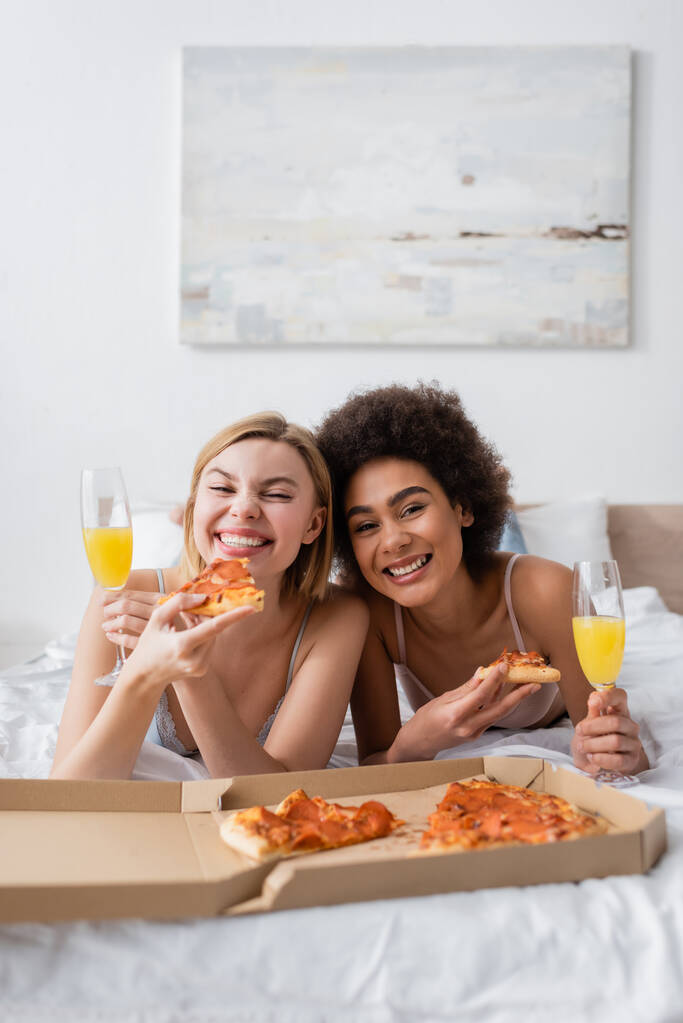 cheerful interracial friends with tasty pizza and cocktails in champagne glasses lying on bed and looking at camera - Foto, Imagen