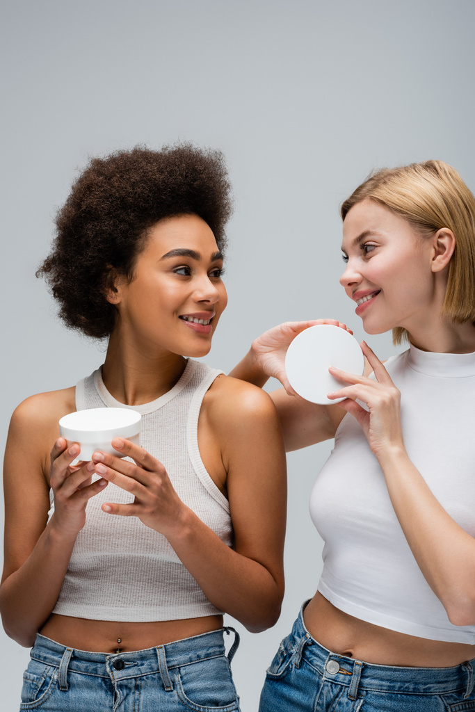 mujeres bastante interracial en tapas blancas sosteniendo contenedores con crema cosmética y sonriéndose aislados en gris - Foto, Imagen