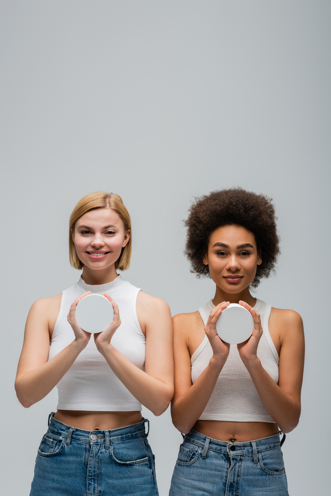 front view of young interracial women wearing white tank tops and holding jars with cosmetic cream isolated on grey - Foto, Bild