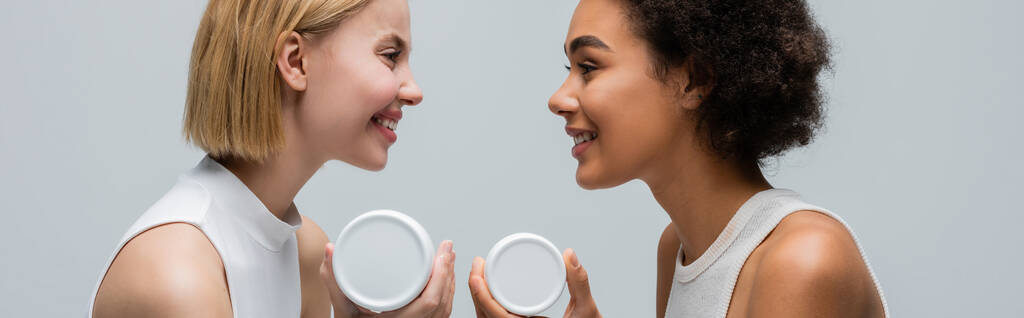 side view of interracial blonde and brunette women with body cream smiling at each other isolated on grey, banner - Fotoğraf, Görsel