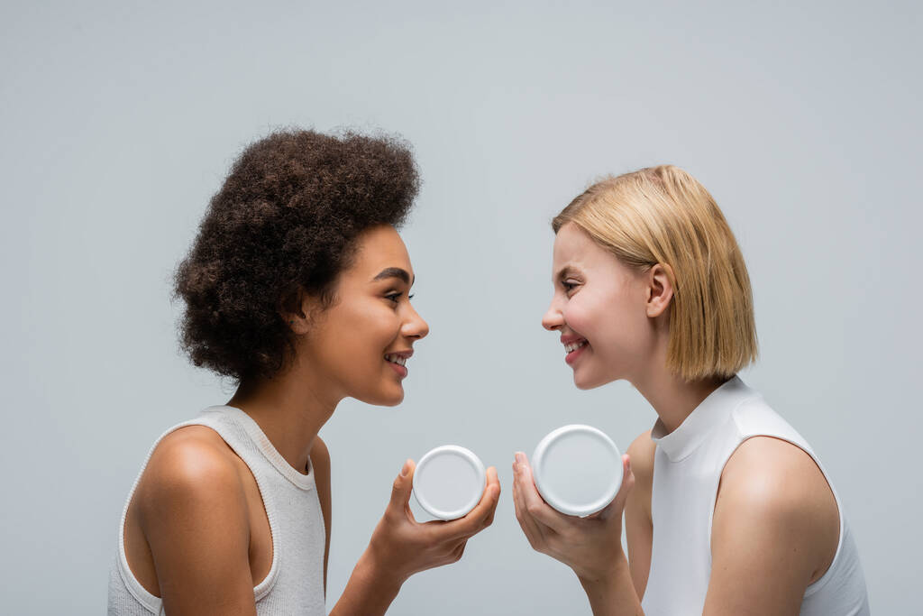 side view of multiethnic blonde and brunette women holding cosmetic cream and smiling at each other isolated on grey - Photo, image