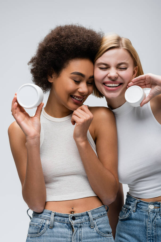 hübsche interrassische Frauen in weißen Tops posieren mit kosmetischer Creme und lächeln mit geschlossenen Augen isoliert auf grau - Foto, Bild