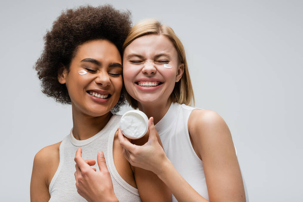 молодые и веселые многоэтнические женщины, улыбающиеся с закрытыми глазами возле банки крема для лица, изолированной на сером - Фото, изображение