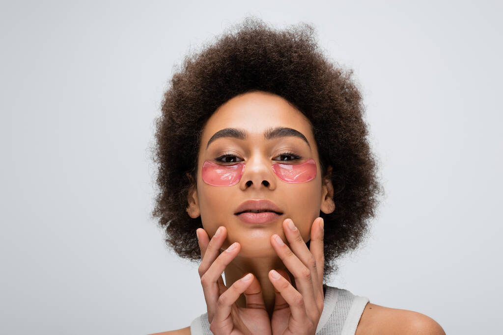curly african american woman touching face with collagen eye patches while looking at camera isolated on grey - Foto, Imagen