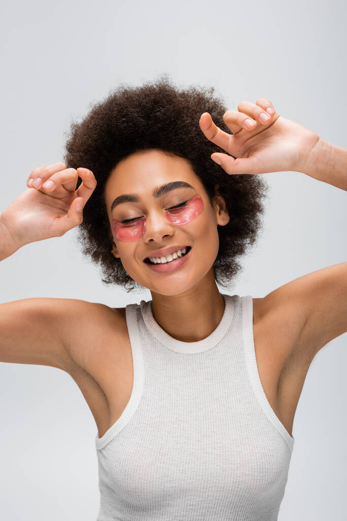 joyful african american woman in white tank top and collagen eye patches posing with closed eyes isolated on grey - Photo, Image